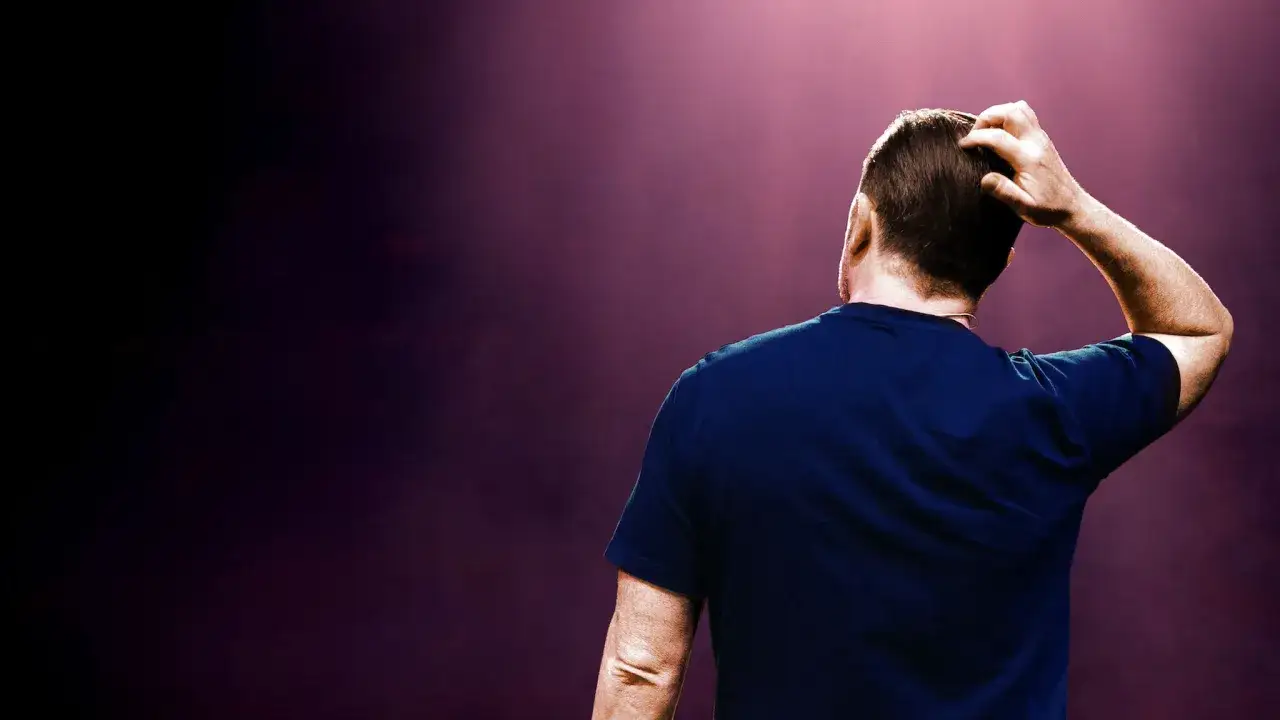 Ricky Gervais stand up, drapiąc się po głowie na tle fioletowego światła.