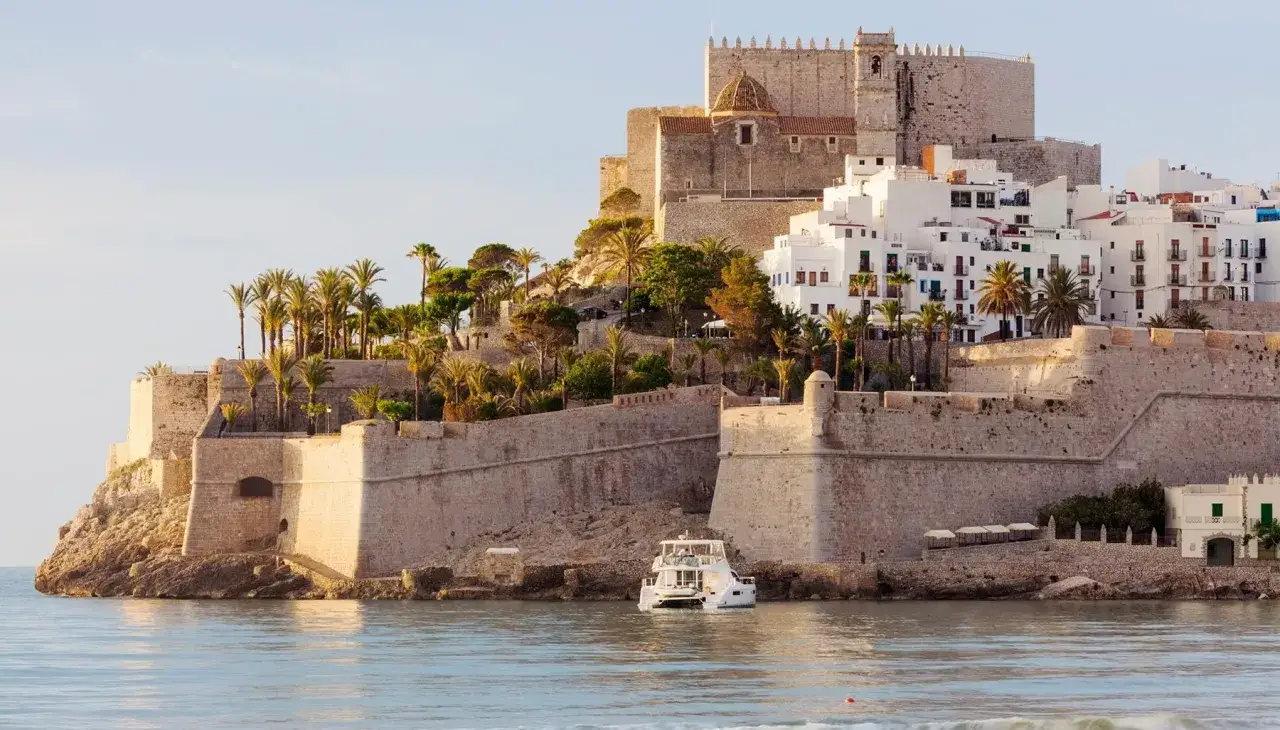 Pe&ntilde;&iacute;scola desde el mar, con su imponente castillo y casas blancas escalonadas en la colina. Un yate blanco navega tranquilamente.