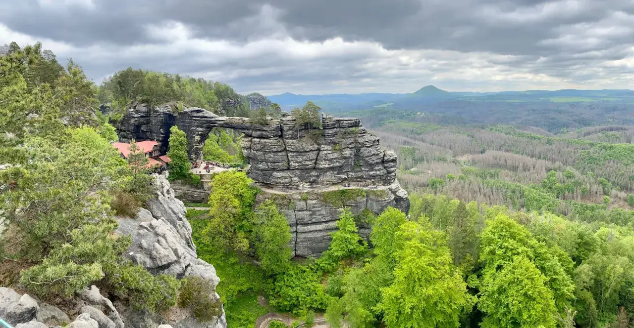 Die Pravcicka brana, ein beeindruckendes Felsentor, ragt &uuml;ber einer gr&uuml;nen Landschaft auf. Ein Gasthaus ist an den Felsen gebaut.