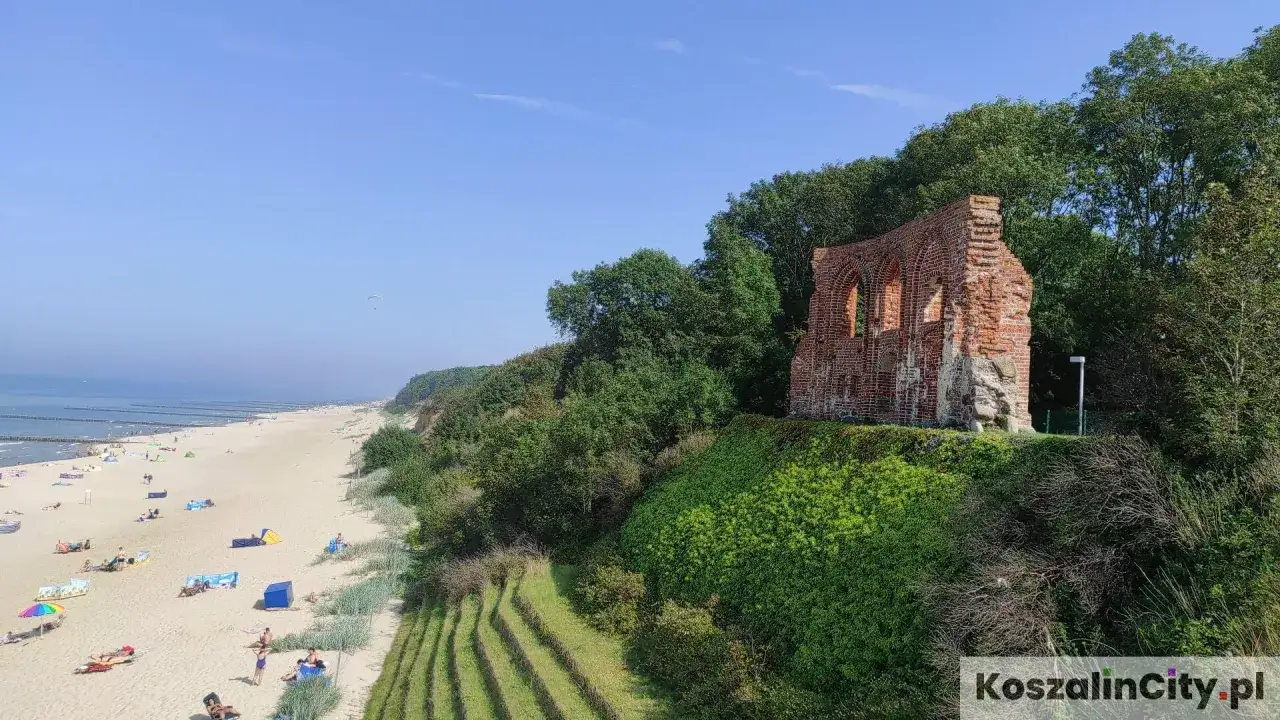 Ruiny zamku w Pobierowie, nad piaszczystą plażą z turystami. W oddali morze i spadochroniarz. To jedne z wielu pobierowo atrakcji.