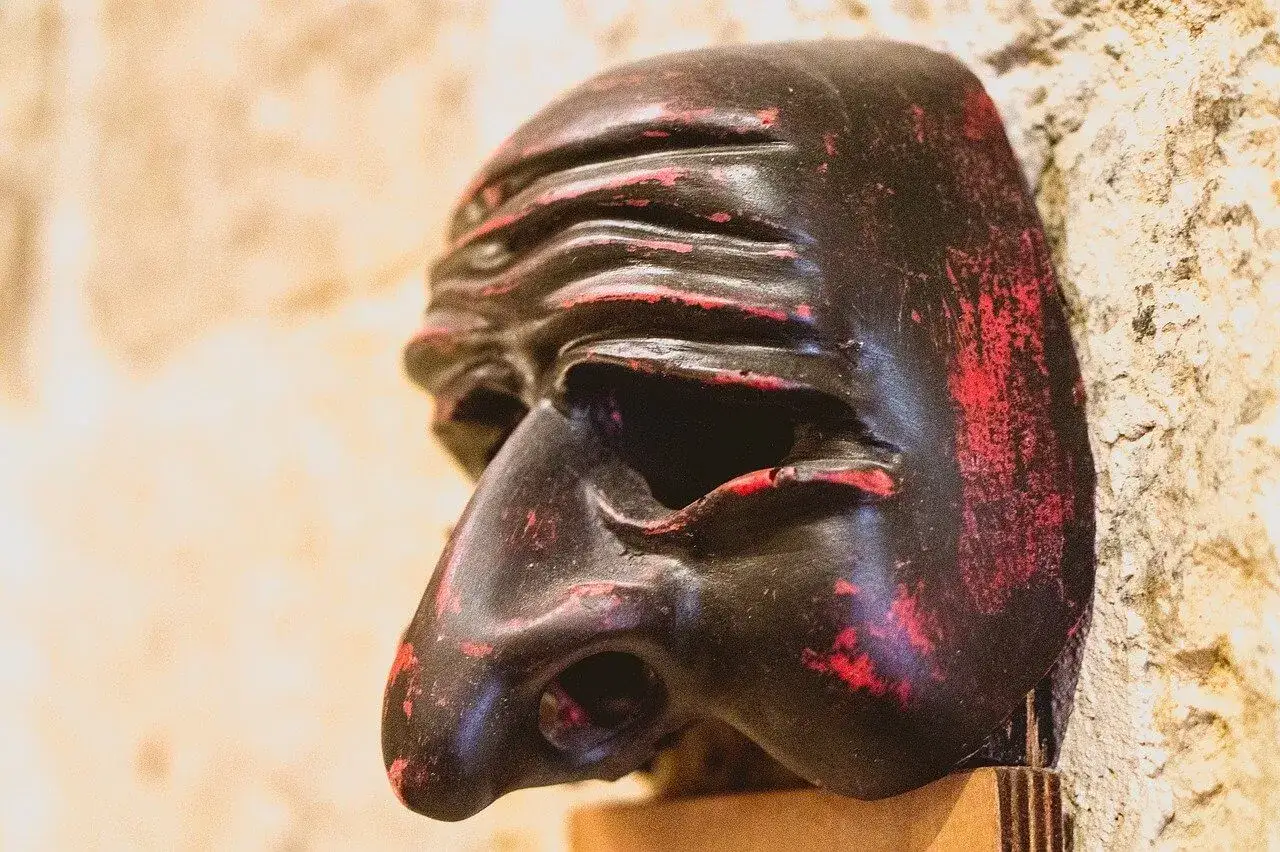 Busto di Pulcinella, maschera nera con dettagli rossi consumati, appoggiata su uno sfondo chiaro e ruvido.