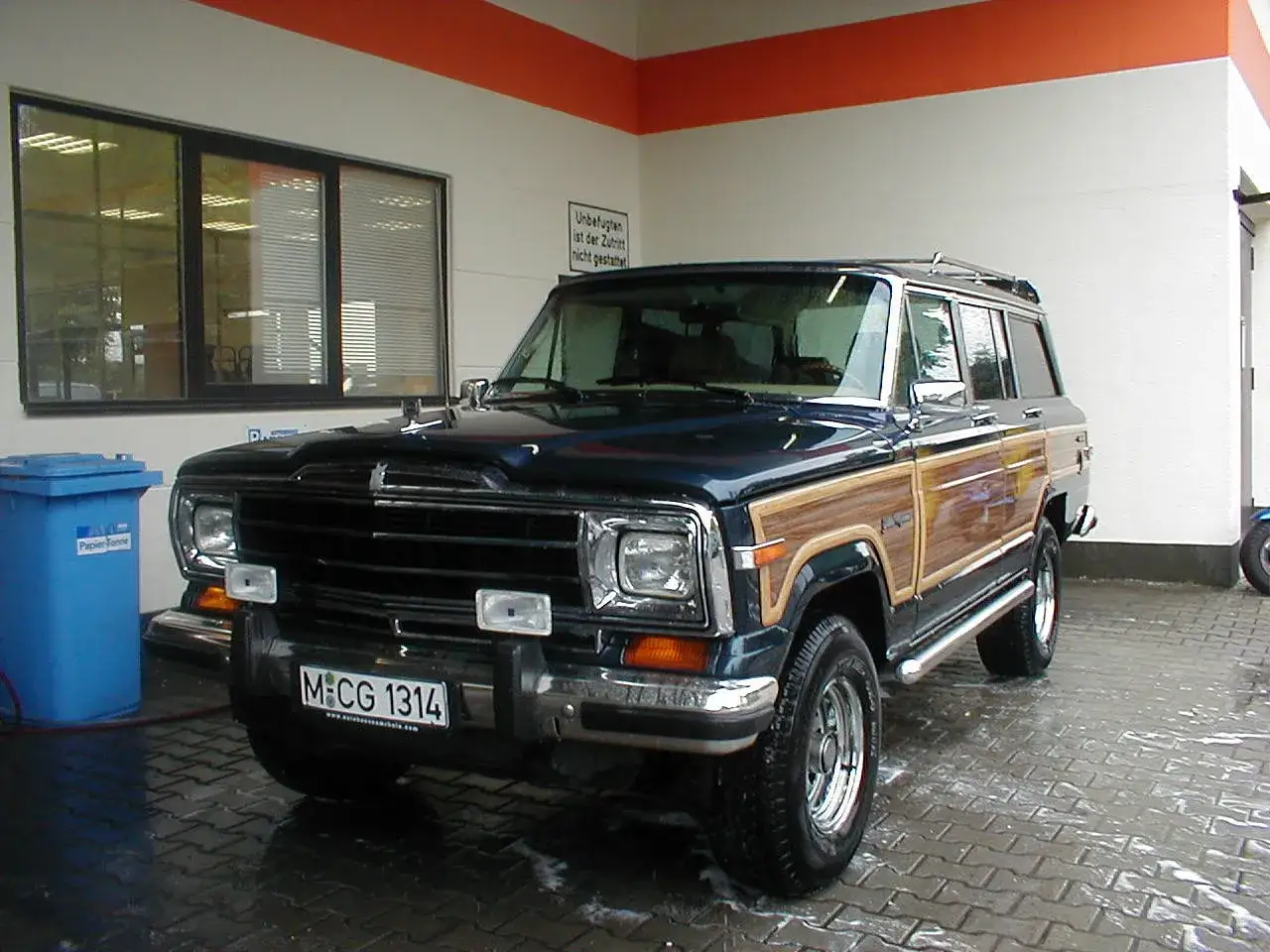 Ein amerikanischer Oldtimer, ein dunkelblauer Jeep Grand Wagoneer mit Holzverkleidung, steht auf nasser Pflasterung.