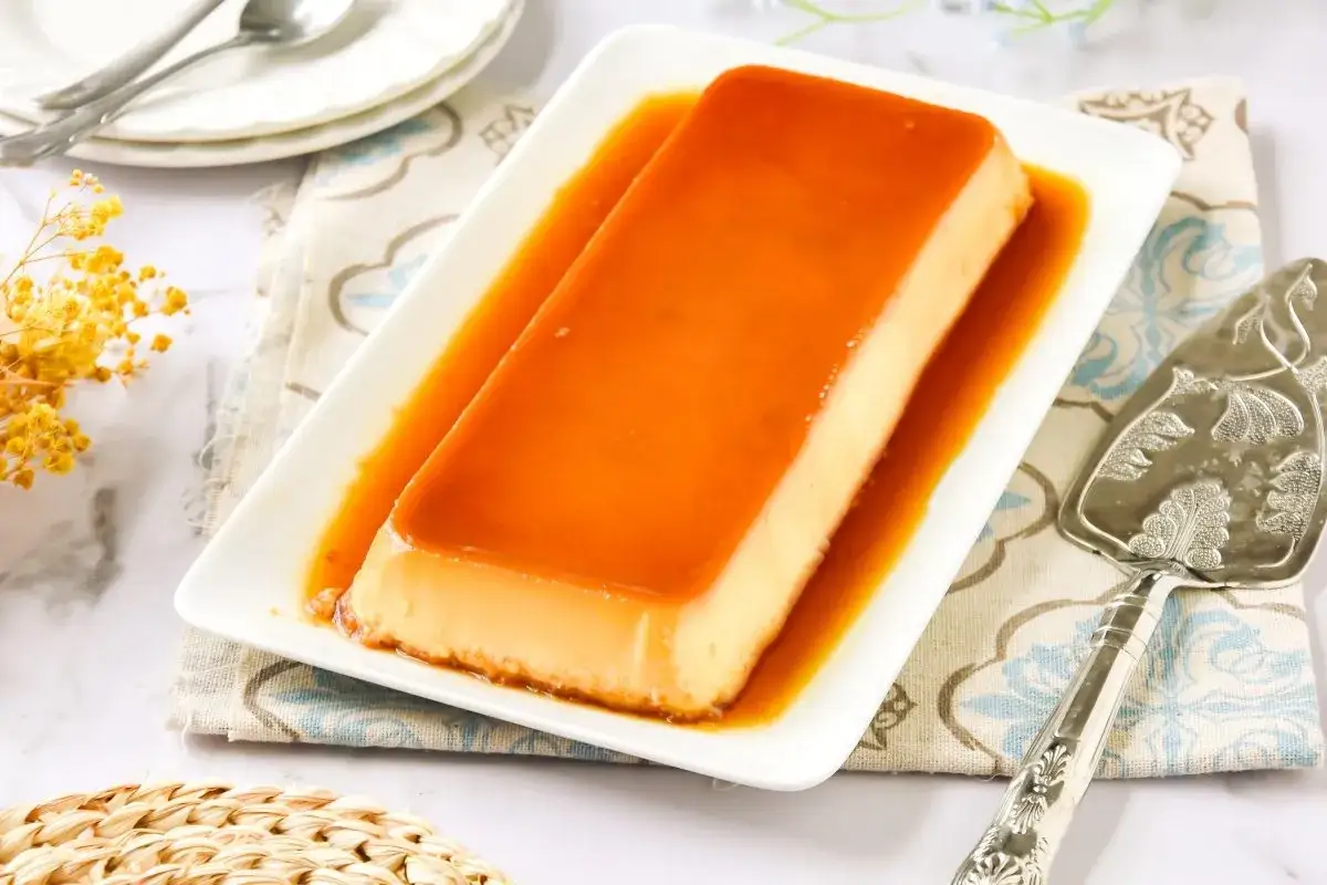 Delicioso flan de huevo casero bañado en caramelo dorado, servido en plato blanco con un elegante utensilio para servir.