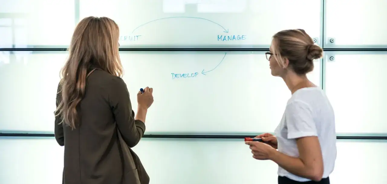 Two women collaborate on a whiteboard, mapping out a cycle of 