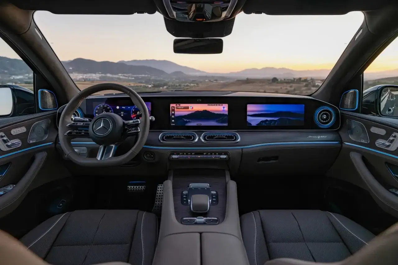 Wnętrze luksusowego **Mercedes-Benz GLS** z podświetleniem ambientowym i dwoma ekranami. Widok na górzysty krajobraz o zachodzie słońca.