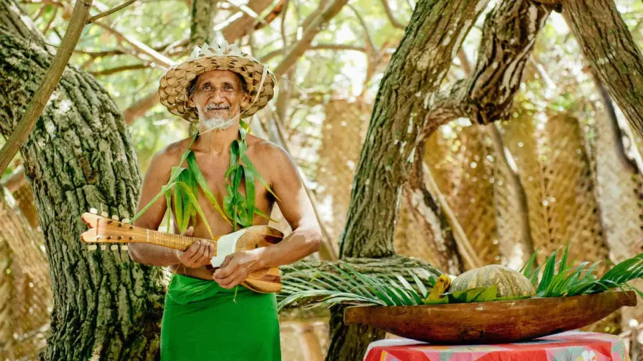 Un homme souriant, v&ecirc;tu d'un pagne vert et d'un chapeau de paille, tient un ukul&eacute;l&eacute;. Il &eacute;voque l'ukul&eacute;l&eacute; origine, un instrument aux racines polyn&eacute;siennes.