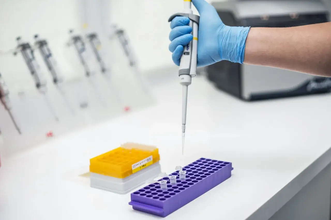 Ręka w niebieskiej rękawiczce trzyma pipetę nad probówkami. W tle stoją kolejne pipety i sprzęt laboratoryjny, sugerując pracę nad analizą, być może badanie kariotypu.