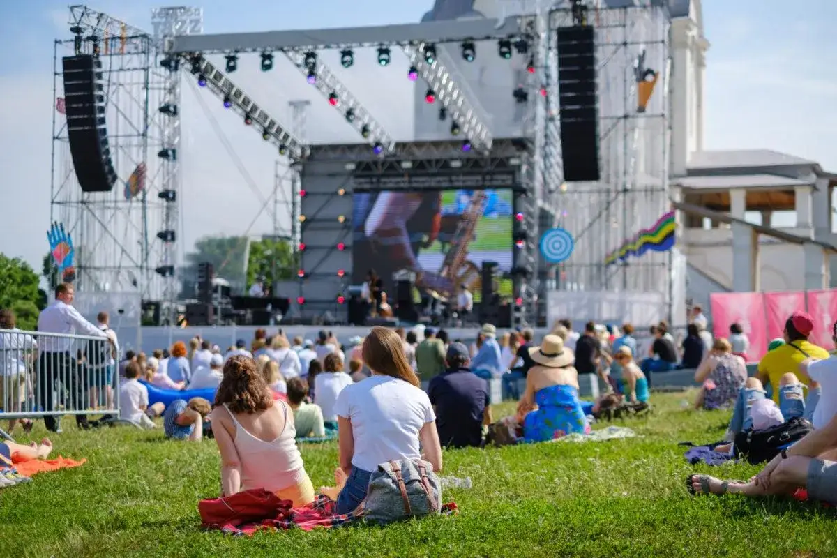 Ludzie siedzą na trawie, oglądając koncert na dużej scenie. To jeden z wielu letnich festiwali w Polsce, pełen muzyki i dobrej zabawy.