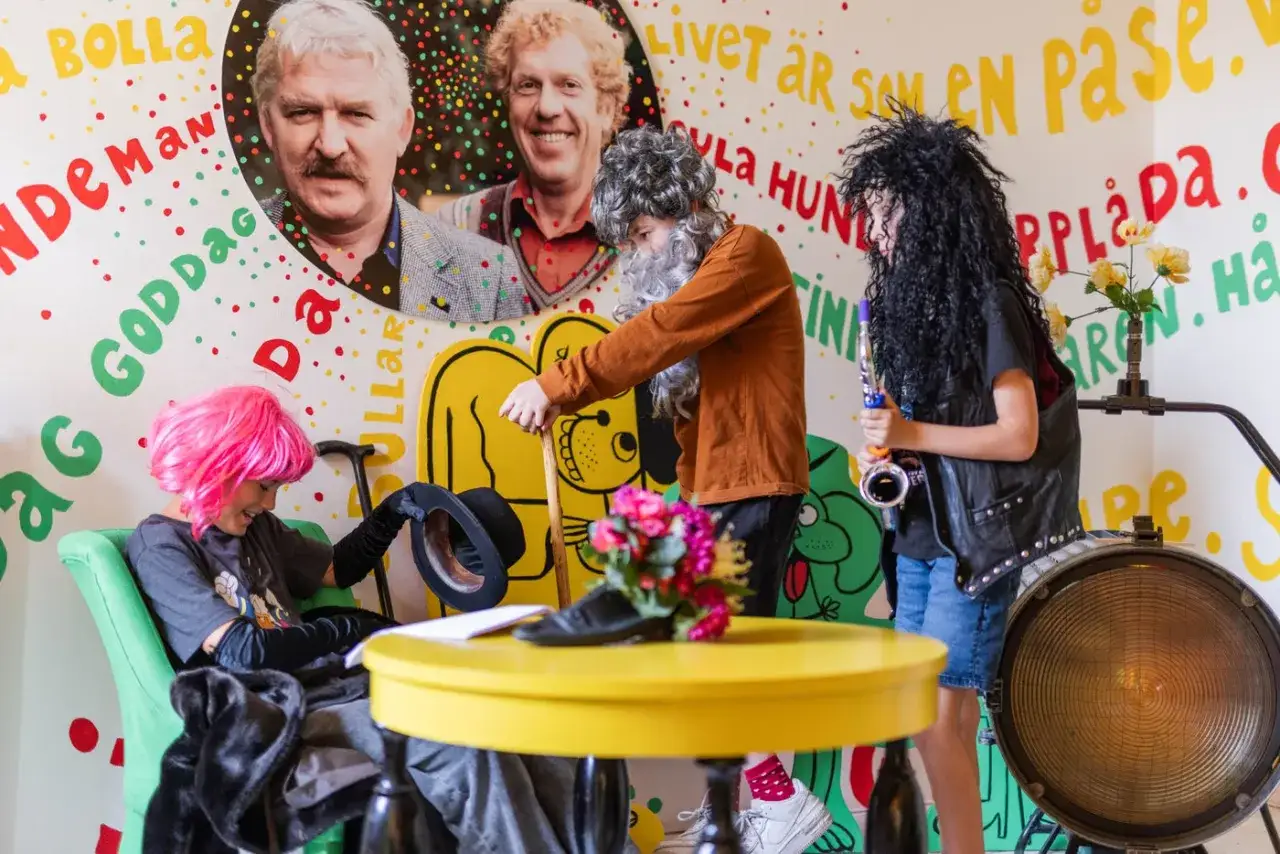 Barn klädda som Eva Rydberg och hennes kollegor leker med hattar och instrument framför en vägg med glada färger och text.
