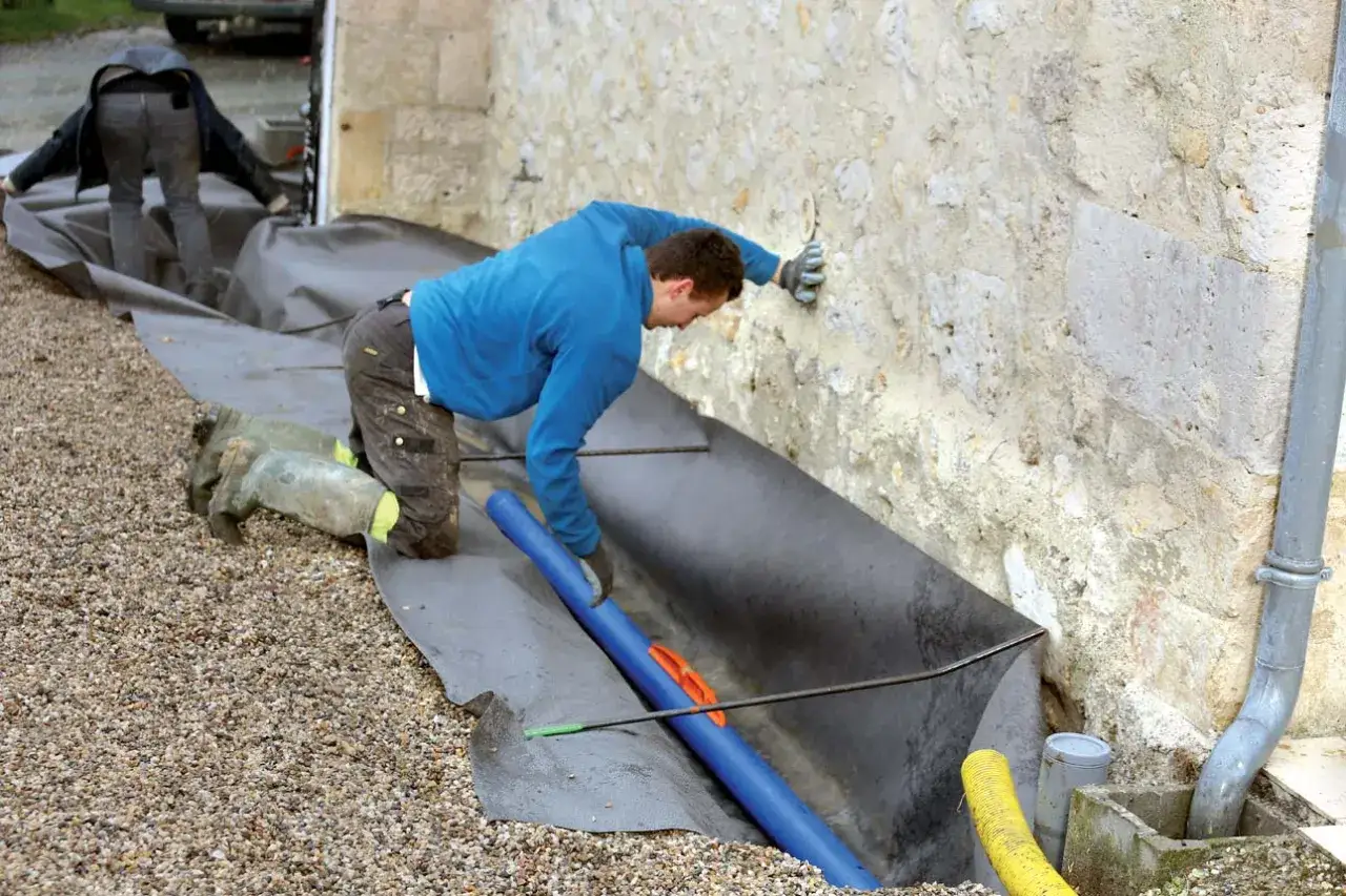 Installation d'un drain de maison. Des ouvriers travaillent à la pose d'un drain autour d'une maison en pierre.