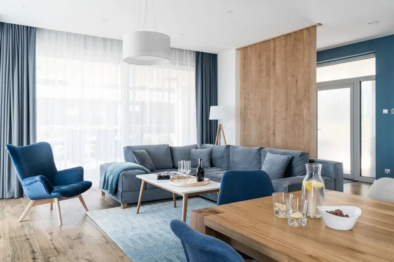 Elegancki salon z sofą i fotelem w odcieniach beżu i granatu. Drewniana ściana dzieli przestrzeń, tworząc przytulną atmosferę.