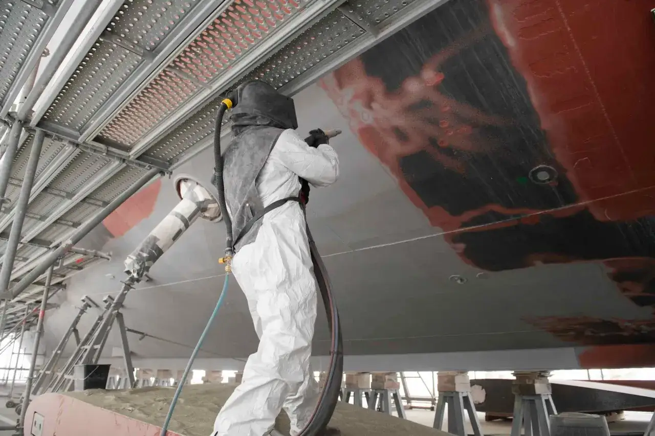 Travailleur en combinaison intégrale sablant la coque d'un bateau pour un traitement antifouling.