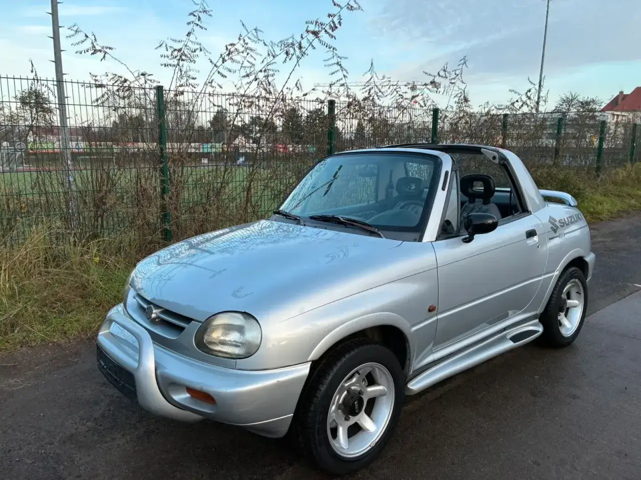 Srebrny Suzuki X-90 z otwartym dachem, zaparkowany przy płocie i boisku sportowym.