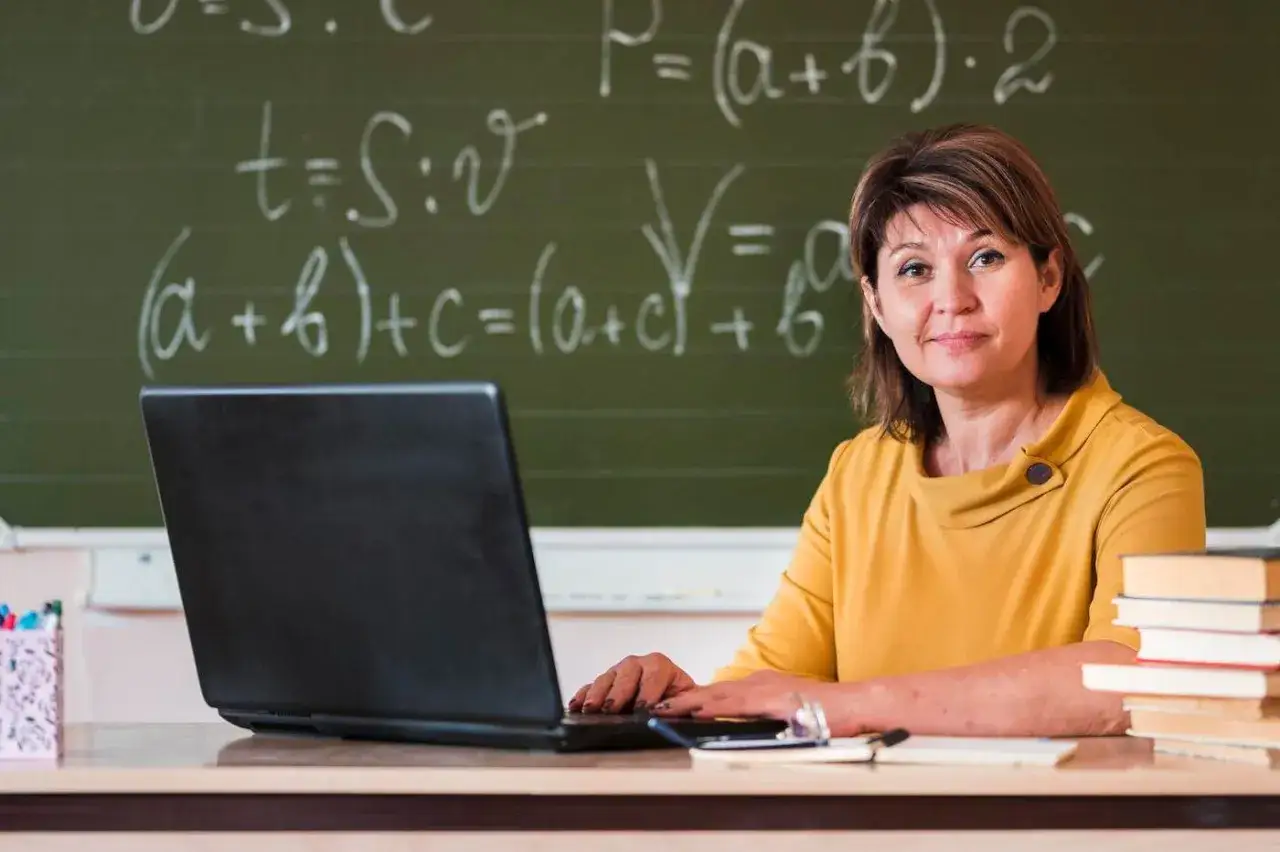 Nauczycielka przy biurku z laptopem i książkami. Na tablicy równania matematyczne. Idealne miejsce na urlop zdrowotny dla kogoś, kto potrzebuje odpoczynku od nauczania.