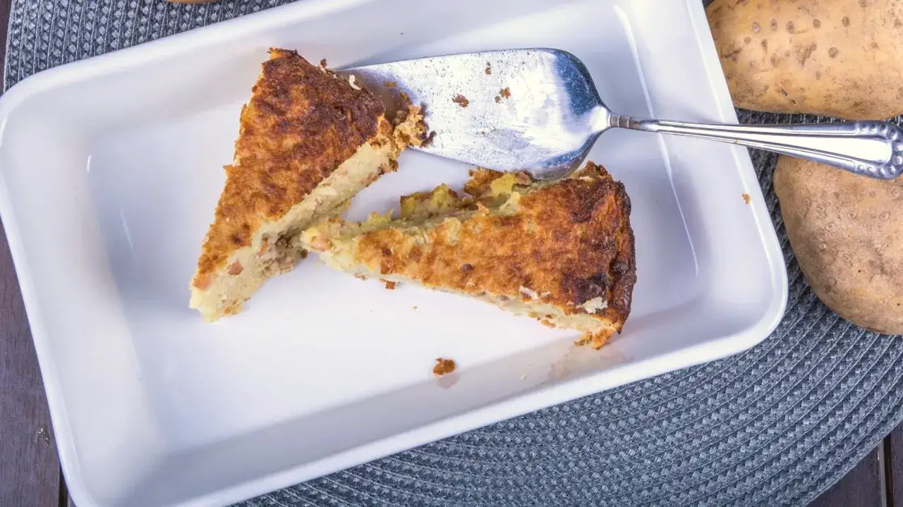 Babka ziemniaczana przepis babci. Dwa kawałki złocistej babki ziemniaczanej w białej brytfance, obok surowe ziemniaki.