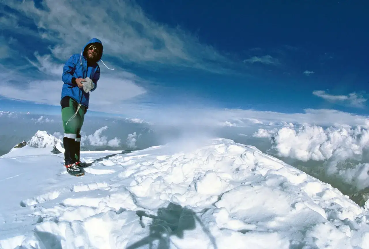 Reinhard Messner, ein Bergsteiger, steht auf einem schneebedeckten Gipfel unter blauem Himmel.