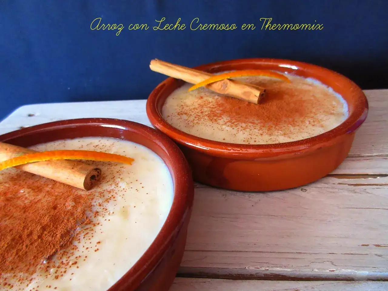 Dos cuencos de barro con cremoso arroz con leche, espolvoreado con canela y decorado con canela en rama y piel de naranja. ¡Perfecto arroz con leche Thermomix!