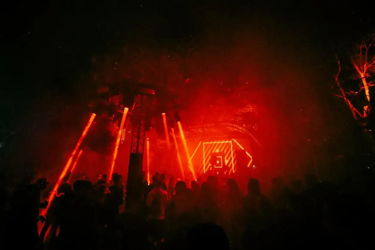Tłumy na festiwalu techno, rozświetlone intensywnymi czerwonymi światłami. W tle widać scenę z neonowym logo.