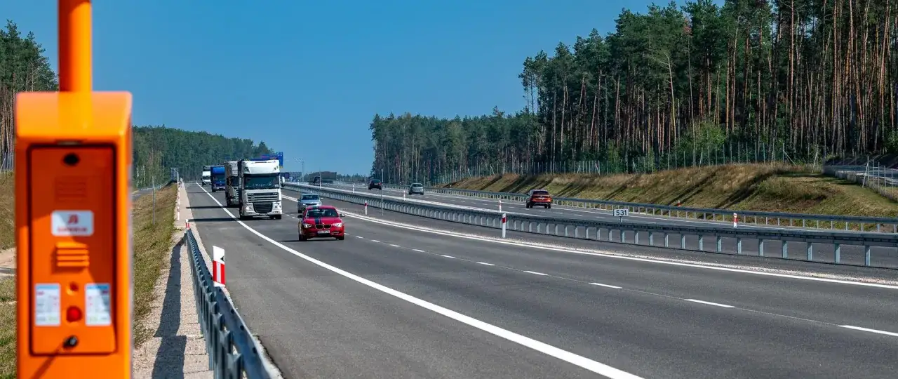 Pomarańczowy terminal przy drodze ekspresowej jednojezdniowej. Samochody osobowe i ciężarowe poruszają się po pustej drodze, otoczonej lasem.
