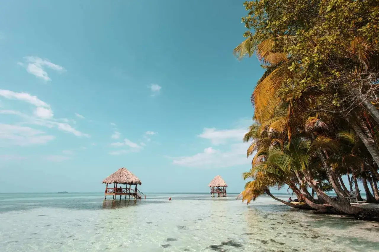 Traumhafte Belize Reisezeit: Zwei Strohhütten im kristallklaren Wasser, Palmen säumen den Strand. Perfekt für Entspannung.