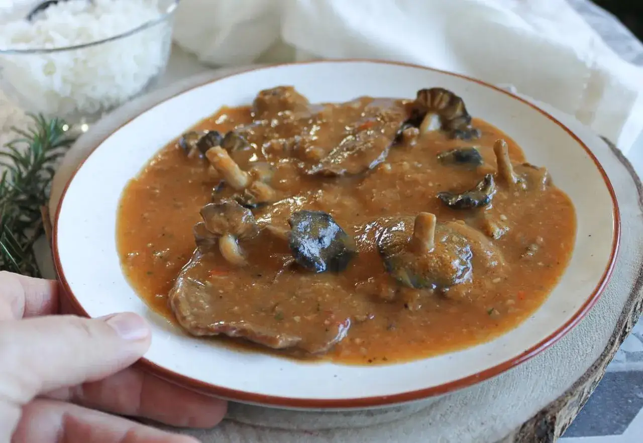 Plato de fricando de ternera con champiñones en salsa. Un tazón de arroz blanco al fondo.