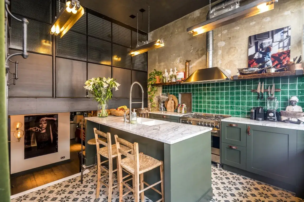 Cocina estilo industrial con isla central de mármol, azulejos verdes y suelo estampado. Lámparas doradas y detalles metálicos completan el ambiente.