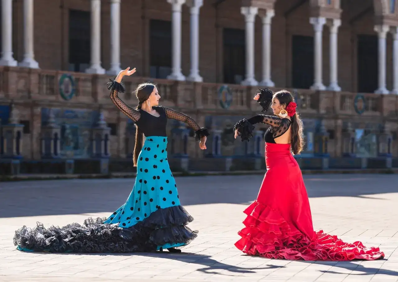 Dos sevillanas flamencas, una con falda turquesa de lunares y otra con falda roja voluminosa, bailan con pasi&oacute;n.