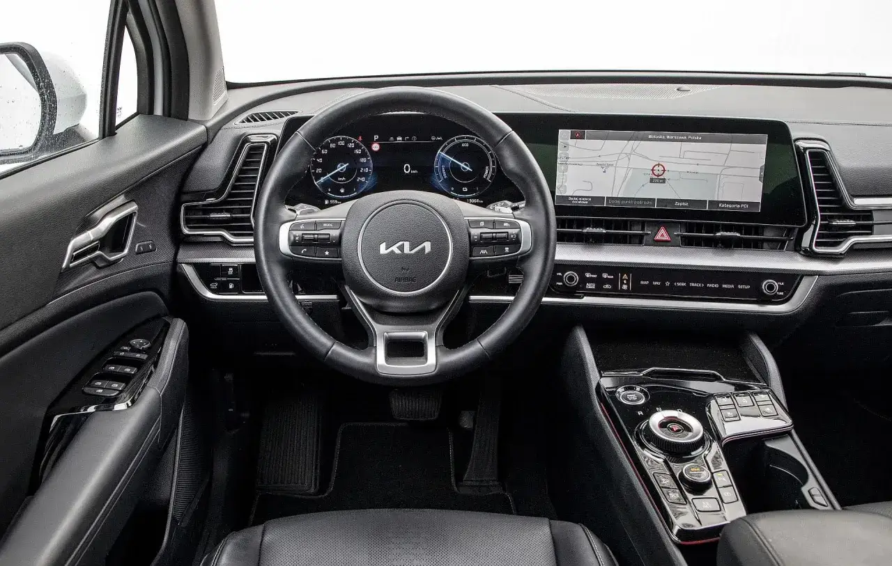 Wnętrze Kia Sportage 1.6 T-GDI: kierownica, cyfrowe zegary i ekran nawigacji z mapą Warszawy.