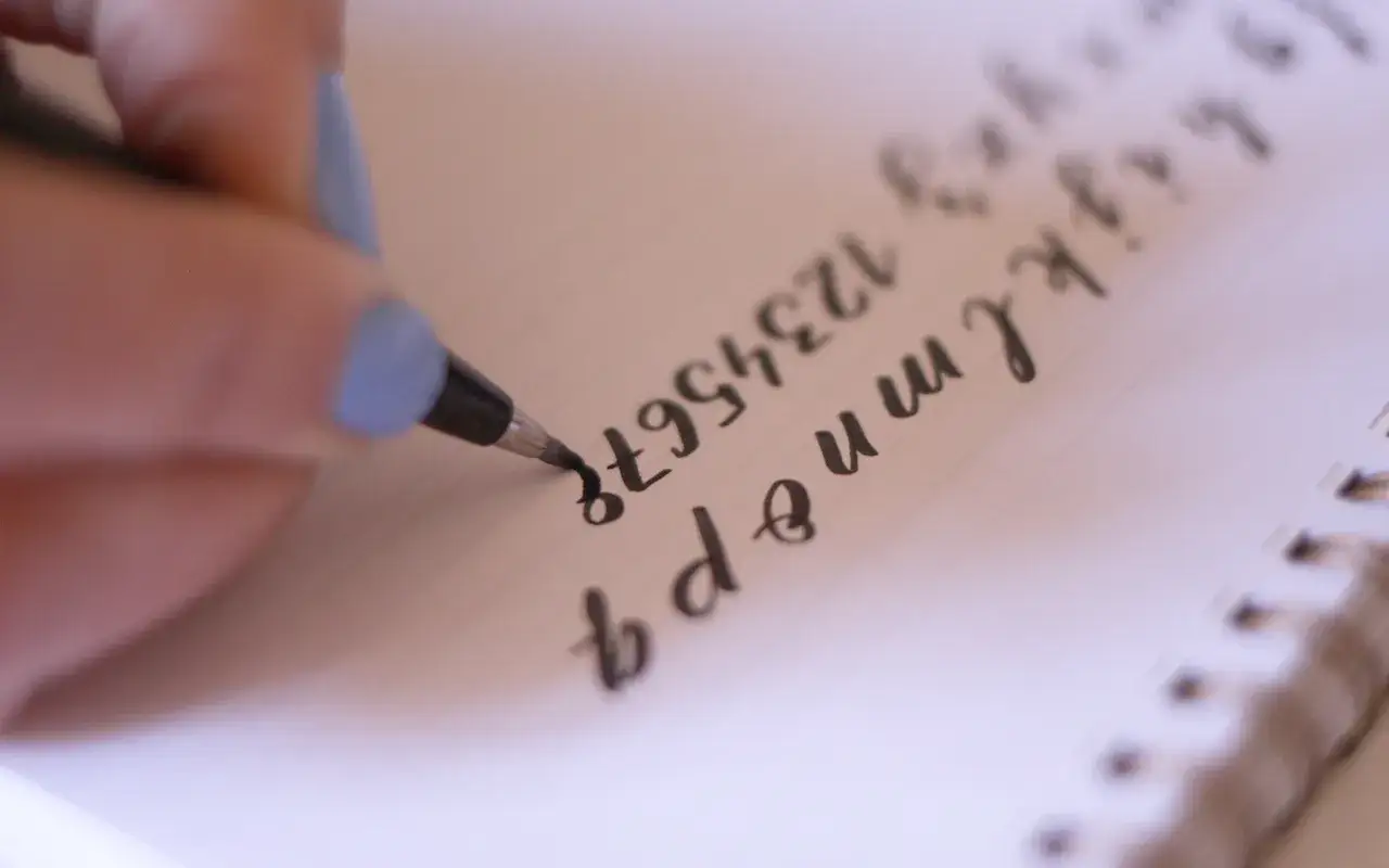 Mano practicando caligrafía inglesa en un cuaderno, trazando letras y números con un rotulador.