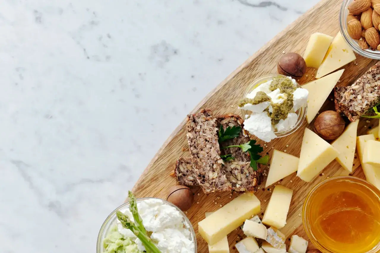 Assortimento di diversi tipi di formaggio, pane integrale, miele, frutta secca e verdure su un tagliere di legno.