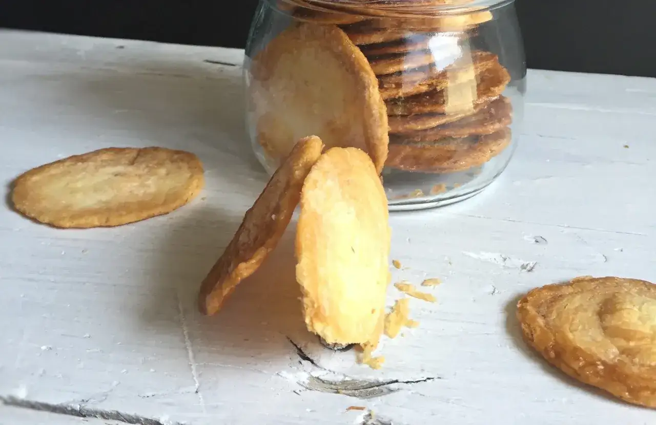 Deliciosas galletas de hojaldre doradas, algunas apiladas en un frasco de vidrio y otras esparcidas sobre una mesa de madera blanca.