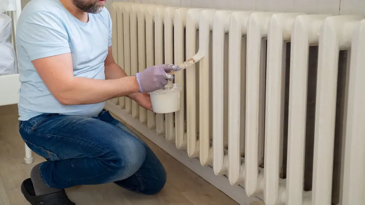 Un homme en jean et t-shirt bleu est à genoux pour peindre un radiateur en fonte avec une peinture blanche.