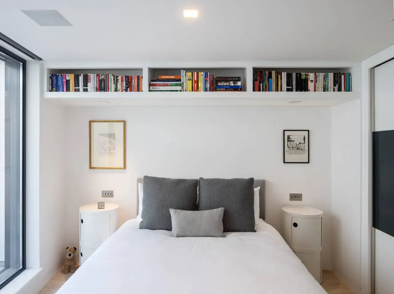 Aménagement petite chambre : lit blanc, tables de nuit rondes, étagères remplies de livres, tableaux et une peluche. Idéal pour optimiser l'espace.