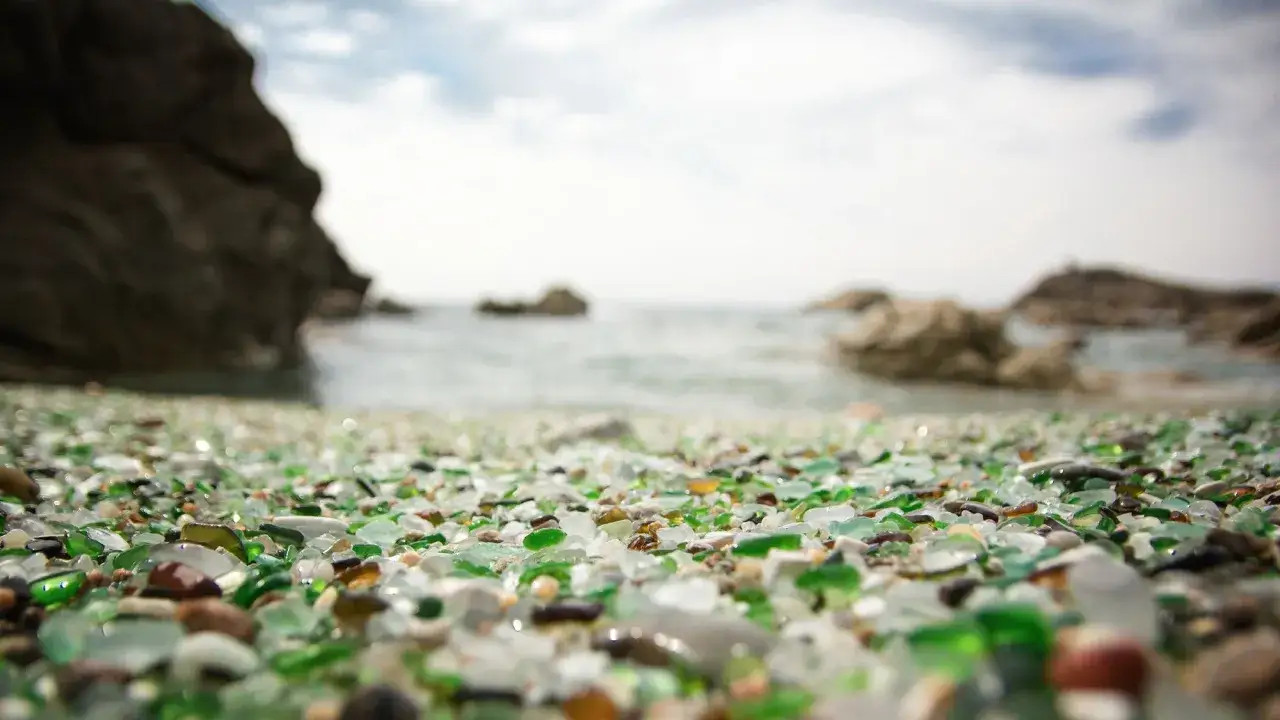 La **playa de los cristales** brilla con fragmentos pulidos por el mar, bajo un cielo nublado y rocas imponentes.