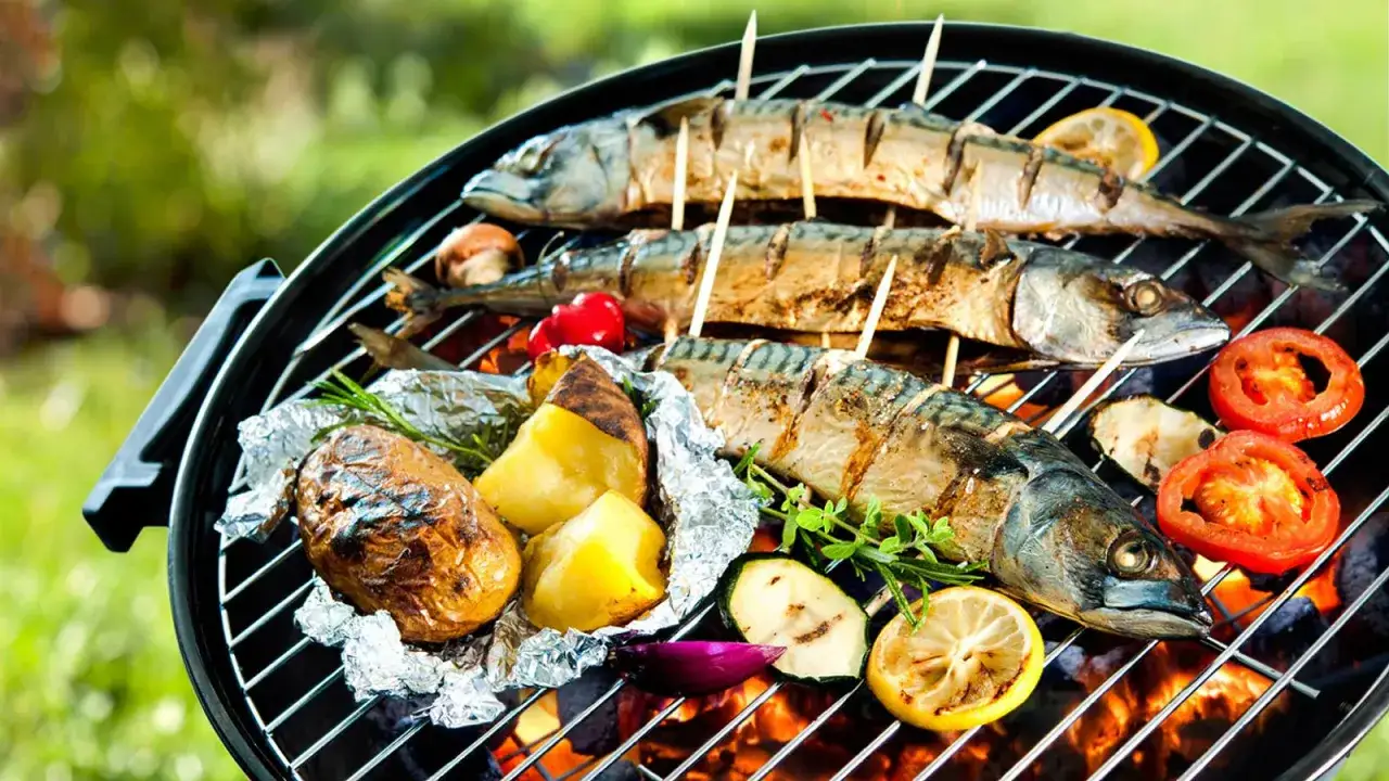 Drei ganze Makrelen werden auf einem Grill zubereitet, zusammen mit Kartoffeln in Alufolie, Tomaten, Zucchini und Zitronenscheiben. Das **Makrele grillen** ist fast fertig.