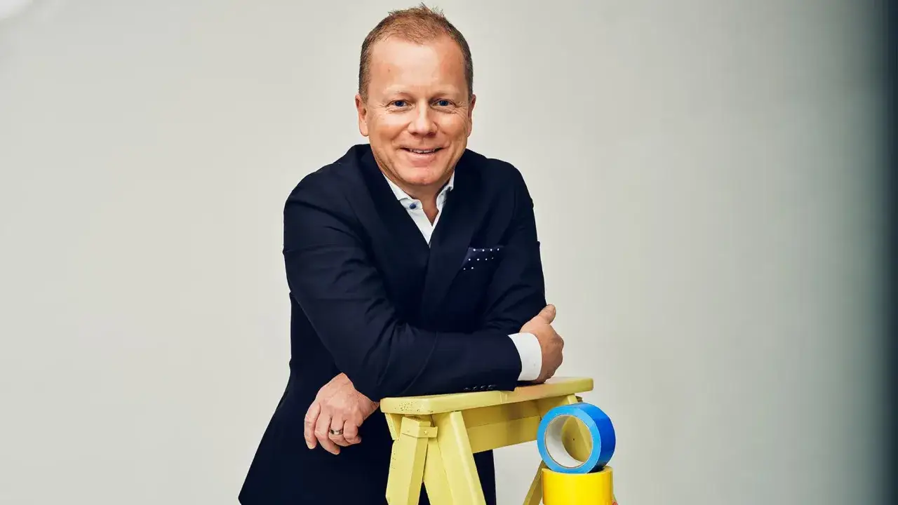 Claes Seldeby, en glad man i kostym, lutar sig mot en gul pall med tejprullar.