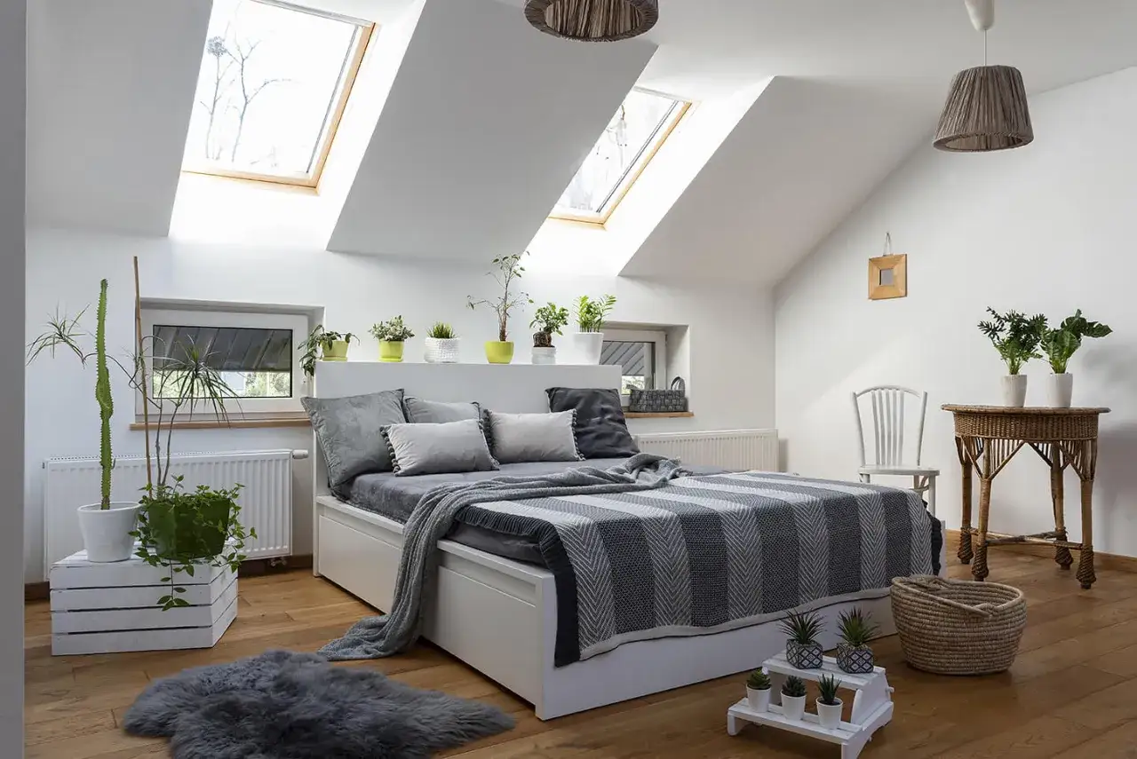 Sypialnia w stylu feng shui: łóżko z szarym pledem, rośliny doniczkowe, drewniana podłoga i okna dachowe tworzą harmonię.
