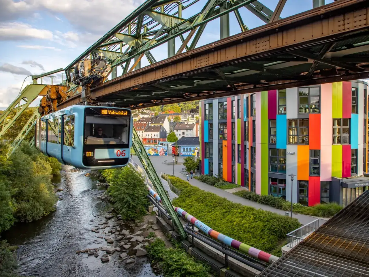 Der Schwebebahnzug f&auml;hrt &uuml;ber einen Fluss, vorbei an einem bunten Geb&auml;ude in Wuppertal.