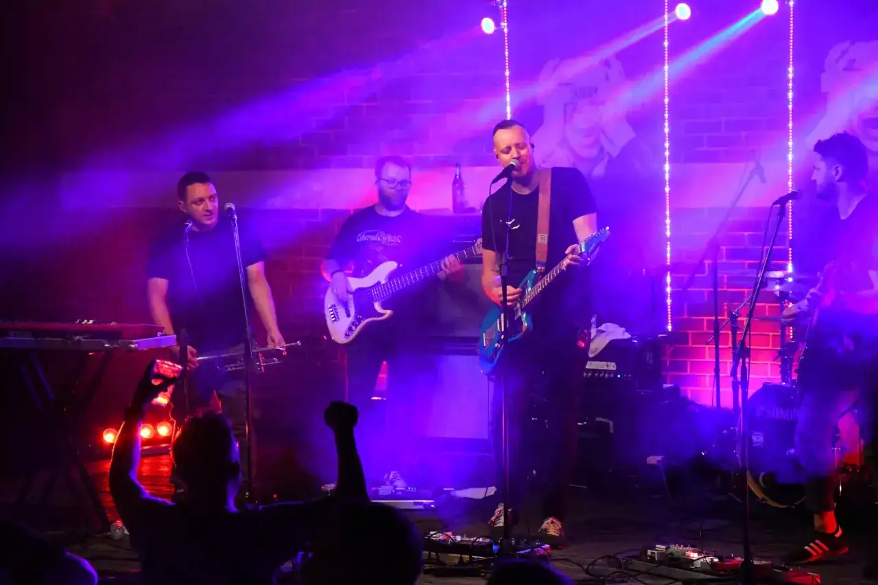 Koniec świata zespół gra na scenie, oświetlony fioletowym światłem. Wokalista śpiewa, gitarzysta basowy i trębacz grają swoje instrumenty.