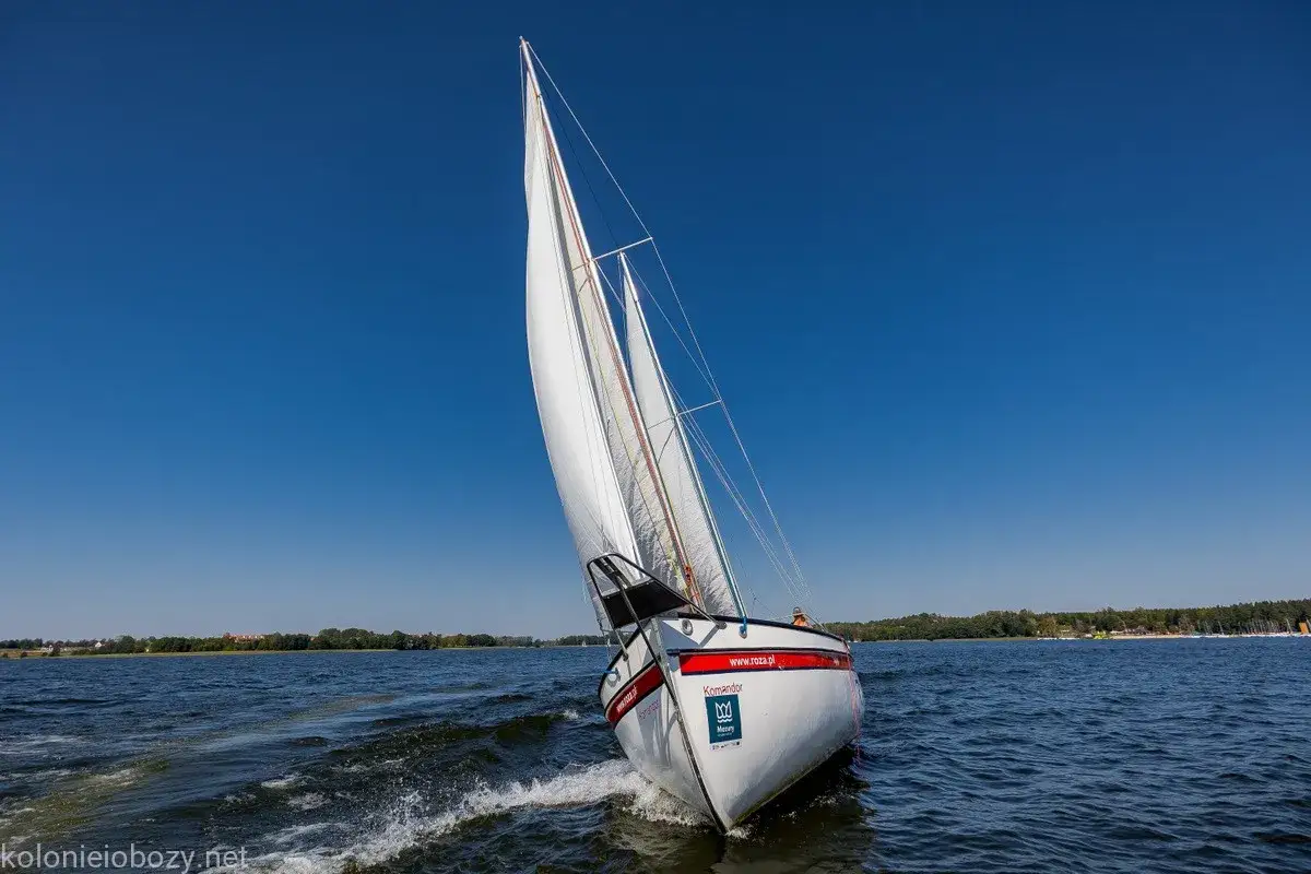Biały jacht z czerwonym pasem pruje fale, idealny na wakacje Mazury. Wiatr napina żagle, tworząc pianę na wodzie.