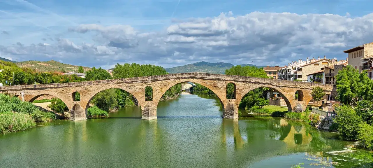 Der historische Puente la Reina &uuml;berspannt mit seinen steinernen B&ouml;gen einen ruhigen Fluss. Dahinter erstreckt sich eine malerische Stadt.