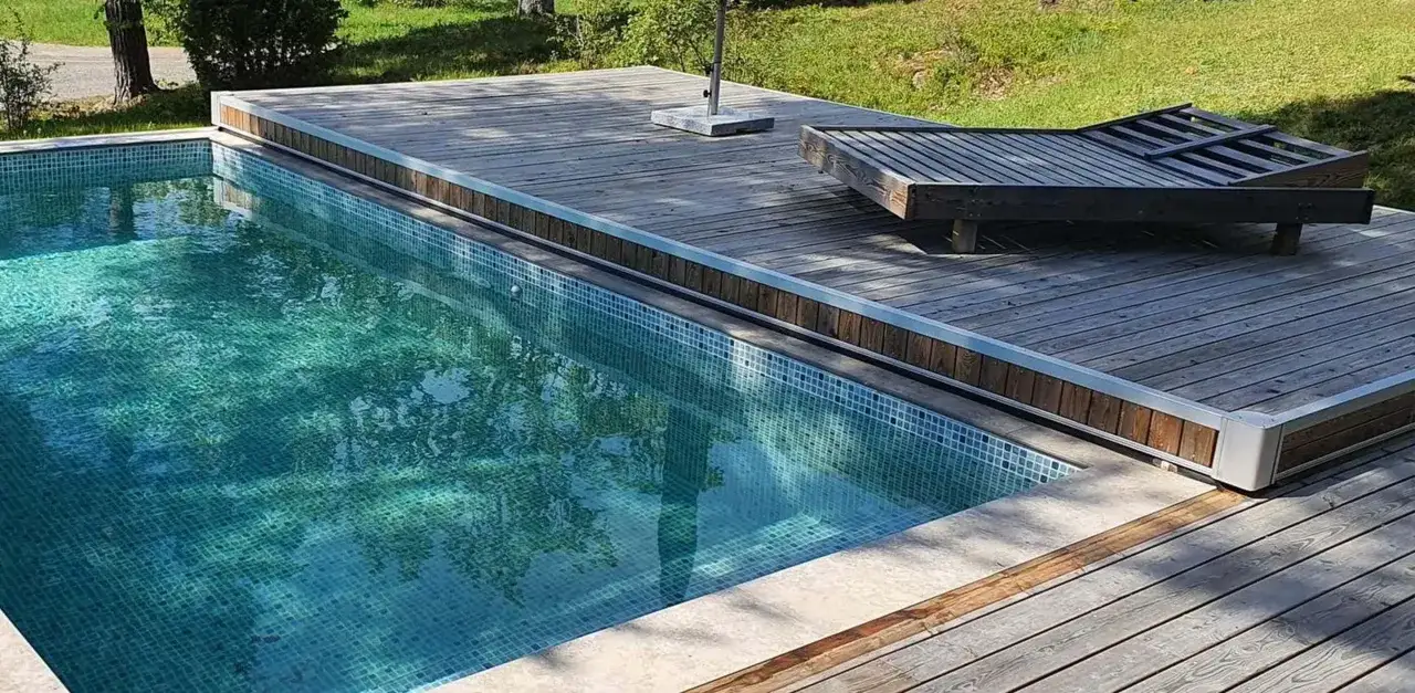 En pool med blå mosaik och en träaltan. En solstol står på altanen, perfekt för att njuta av solen efter att ha byggt eget poolskydd.