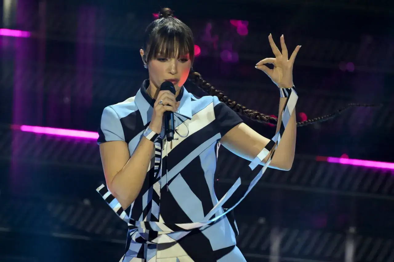 Sara, la cantante, incanta il pubblico con la sua voce e un gesto della mano, indossando un abito geometrico su un palco illuminato.