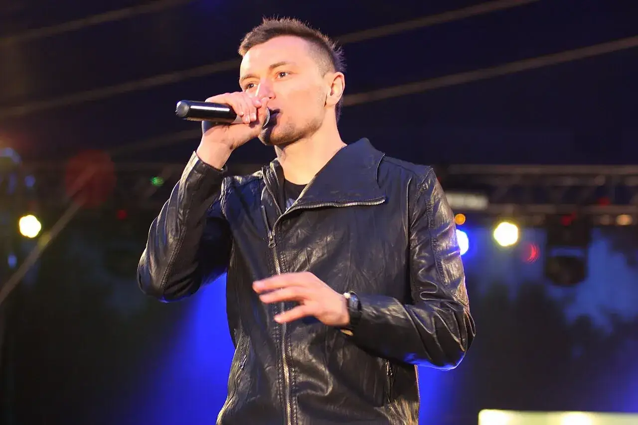 Liber śpiewa na scenie w skórzanej kurtce, oświetlony niebieskim światłem.