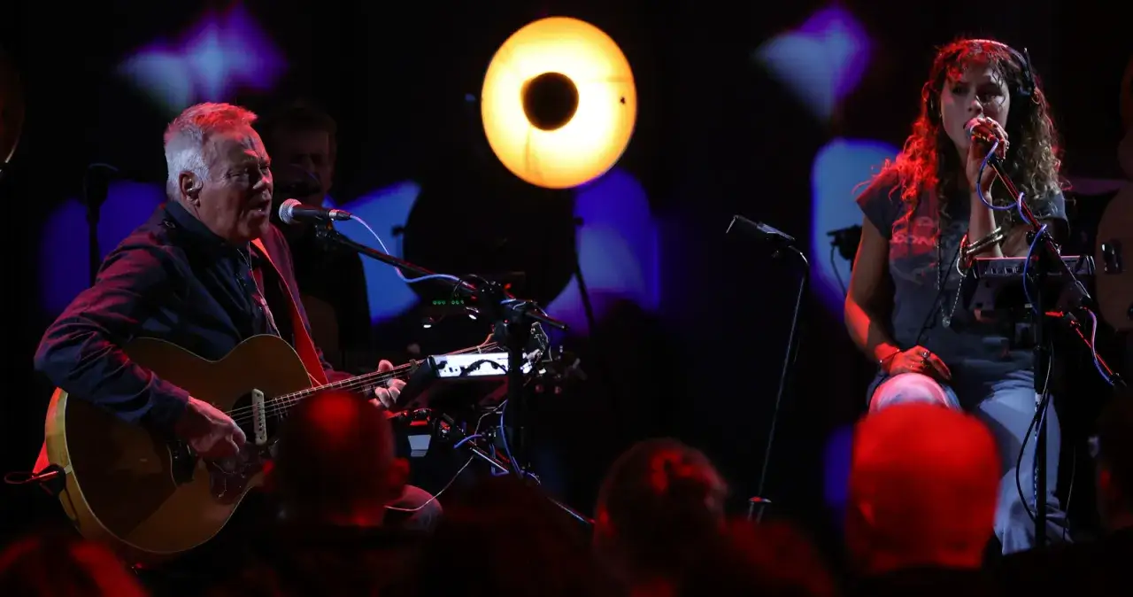 Mężczyzna gra na gitarze, a kobieta śpiewa. Scena oświetlona jest ciepłym, **red tune** światłem.