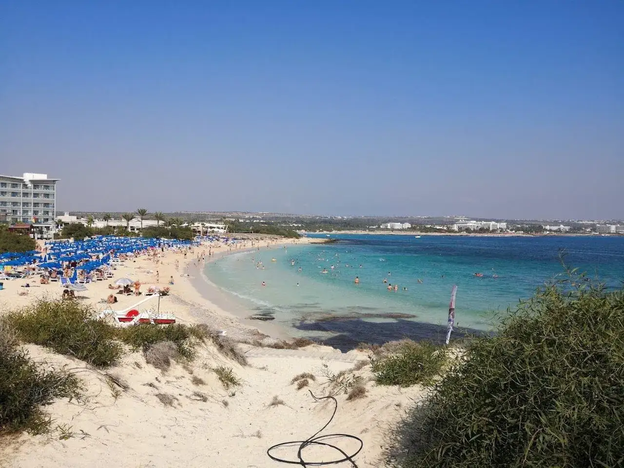 Słoneczny dzień na plaży Makronissos. Tłumy ludzi odpoczywają pod niebieskimi parasolami, kąpią się w turkusowym morzu.
