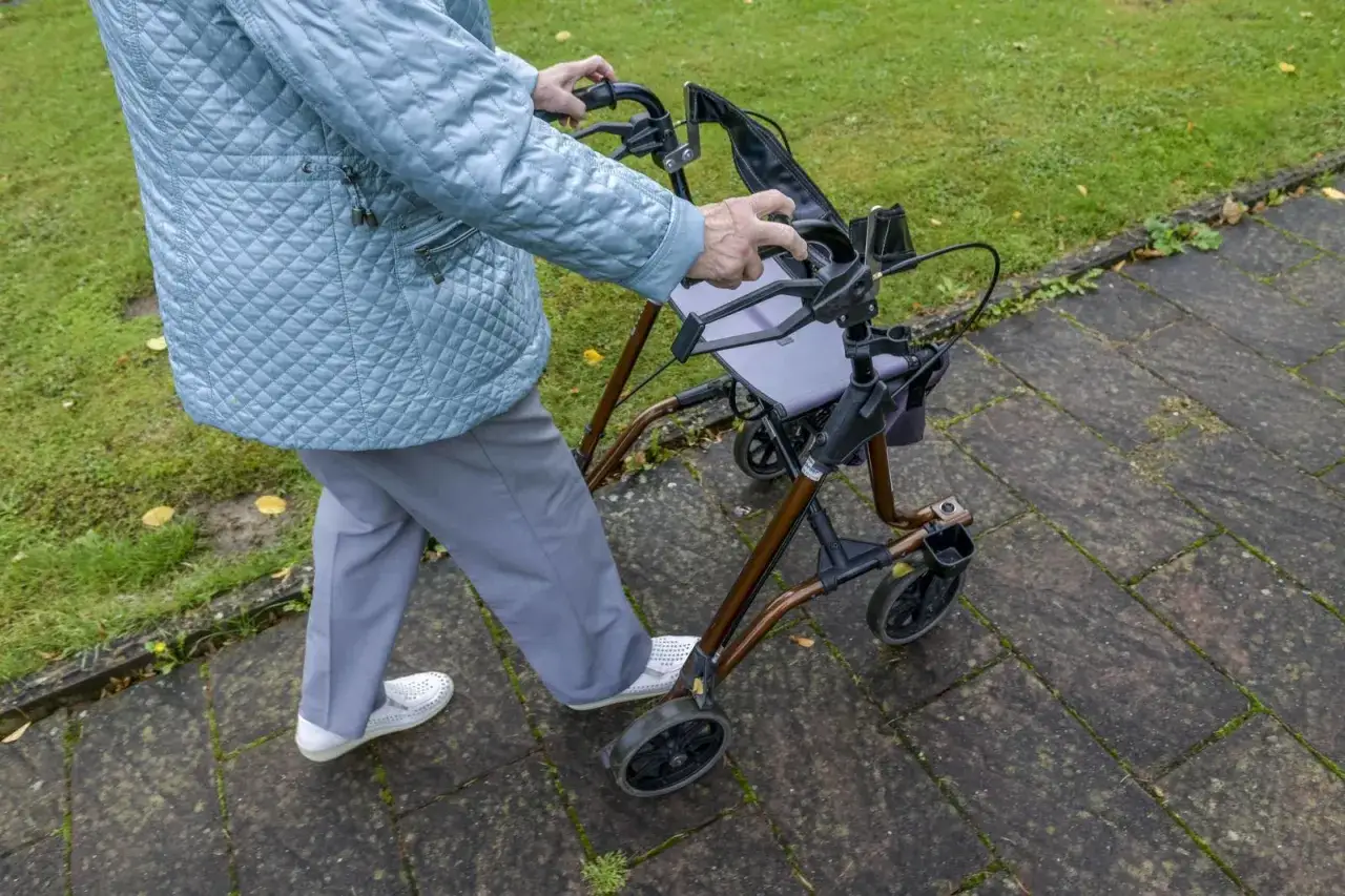 &Auml;ltere Person mit Rollator auf Rezept, Eigentum. Geht auf gepflastertem Weg.