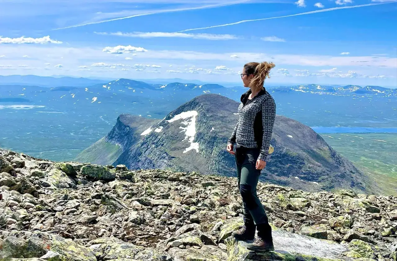 Kvinna p&aring; stenig topp med vidstr&auml;ckt utsikt &ouml;ver fj&auml;llandskap. En karta &ouml;ver Helags syns i bakgrunden.