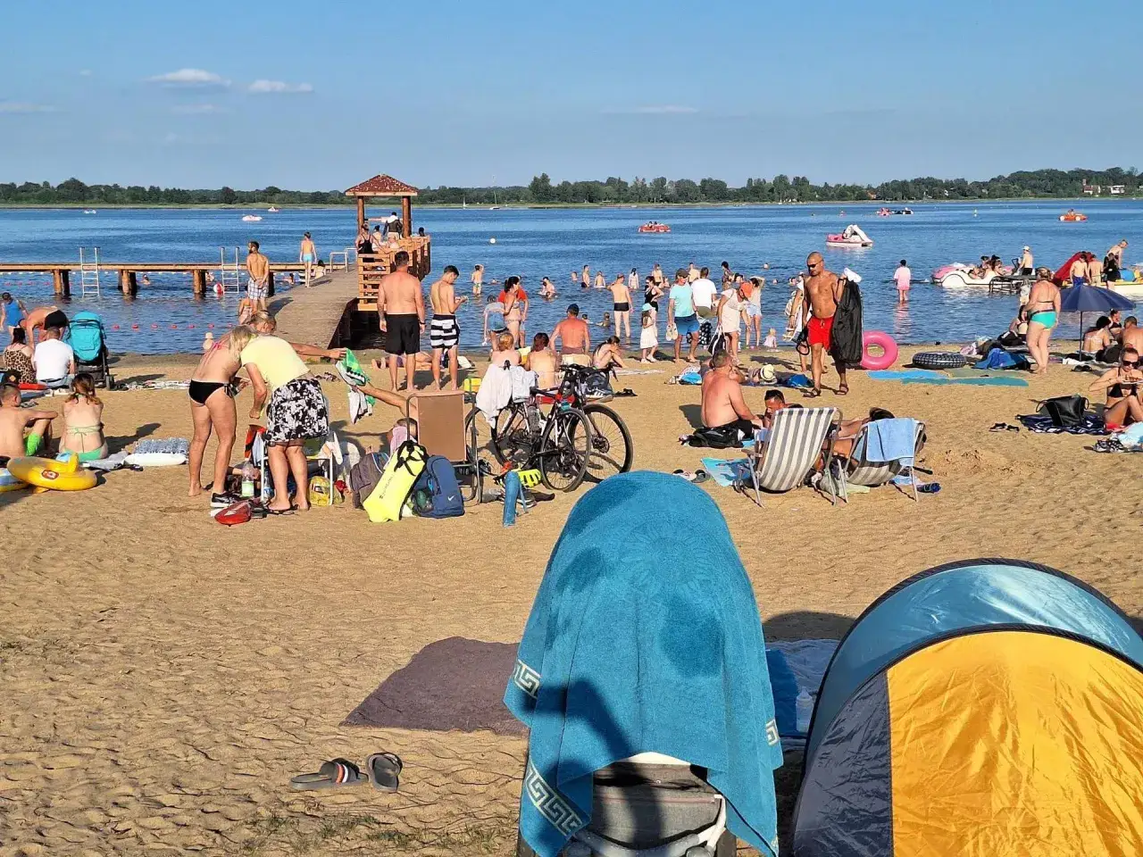 Tłum ludzi na plaży, kąpielisko z pomostem, ł&oacute;dki i rowery wodne. Idealne miejsce, gdzie warto pojechać nad jezioro w Polsce na wakacje.