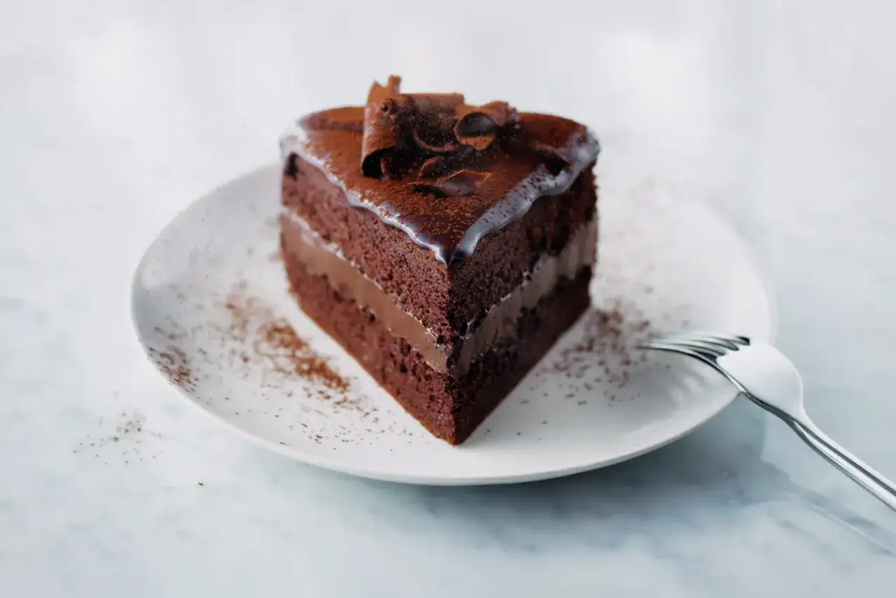 Une part de **sponge cake chocolat** onctueuse, garnie de crème et décorée de copeaux de chocolat, sur une assiette saupoudrée de cacao.